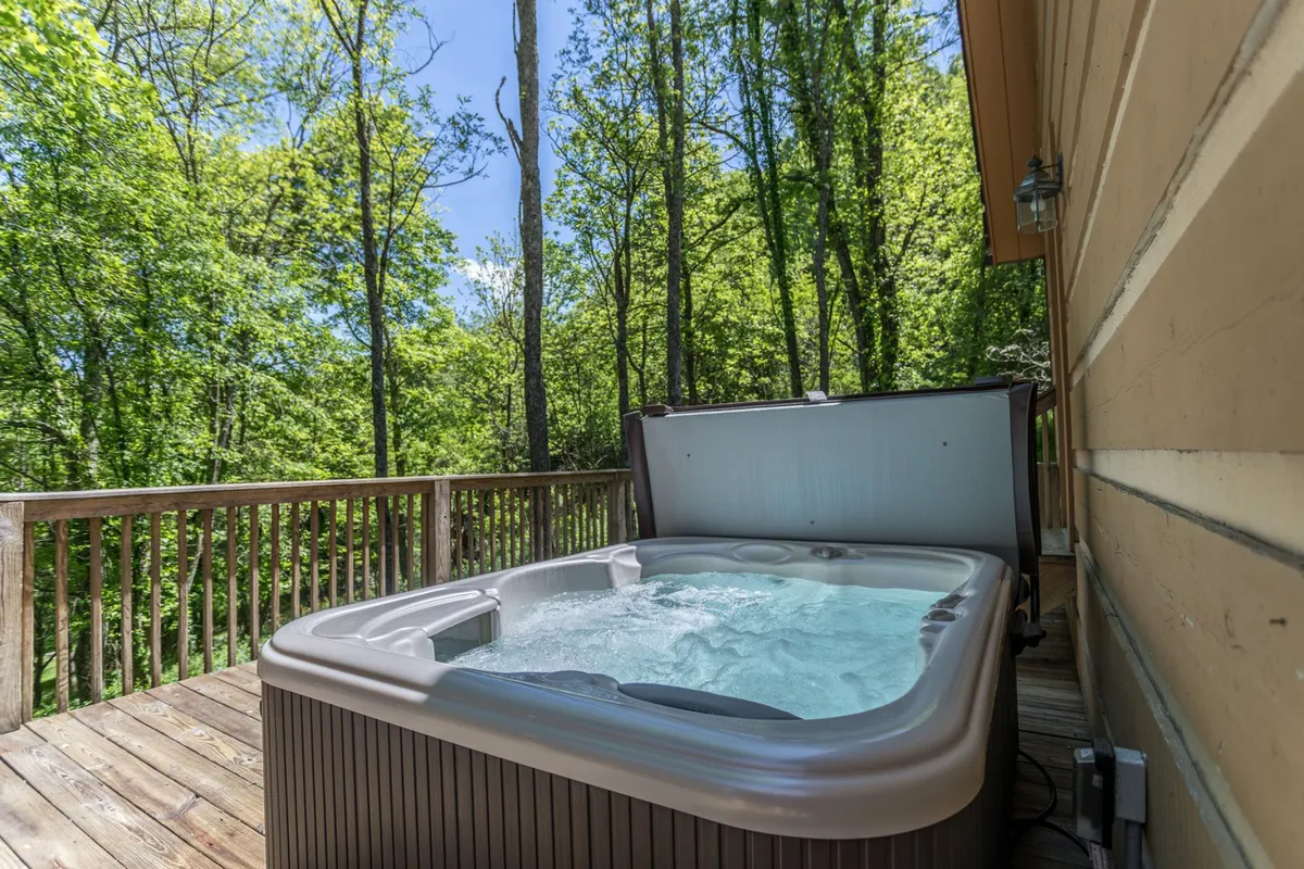 A view of the wooden deck with a bubbling hot tub set against a backdrop of trees, perfect for relaxing, entertaining, and taking in sunset or night sky views. The scene evokes an atmosphere of relaxation and togetherness.