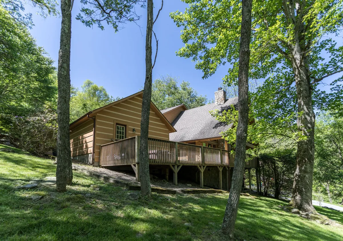 An exterior perspective of the cabin’s wooden deck and stone chimney, encircled by lush trees. The deck invites relaxation and offers a smooth transition between indoor and outdoor living, hinting at easy access from the main living spaces.