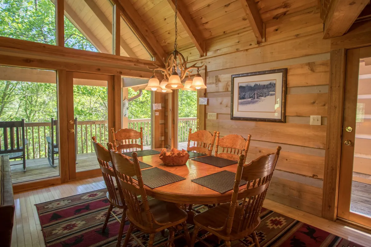 A warm dining nook with a wooden table beneath a charming chandelier, surrounded by large windows and wooden walls. The space feels intimate yet open, with glimpses of the outdoors and a cozy rug enhancing the homely vibe.