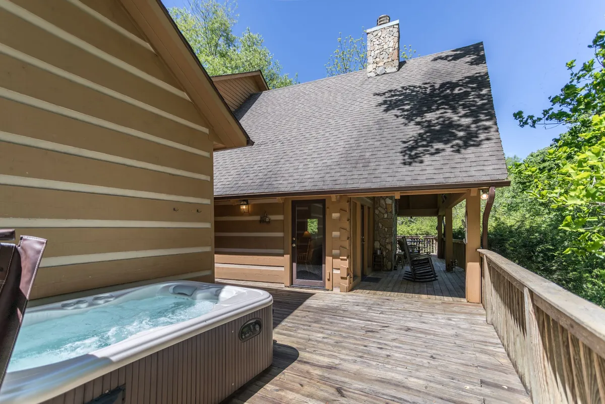An exterior view of a wooden deck featuring a private hot tub, stone chimney, and surrounded by mature trees. The setting is tranquil, perfect for relaxing soaks while enjoying sunset views or stargazing after a day of local adventures.