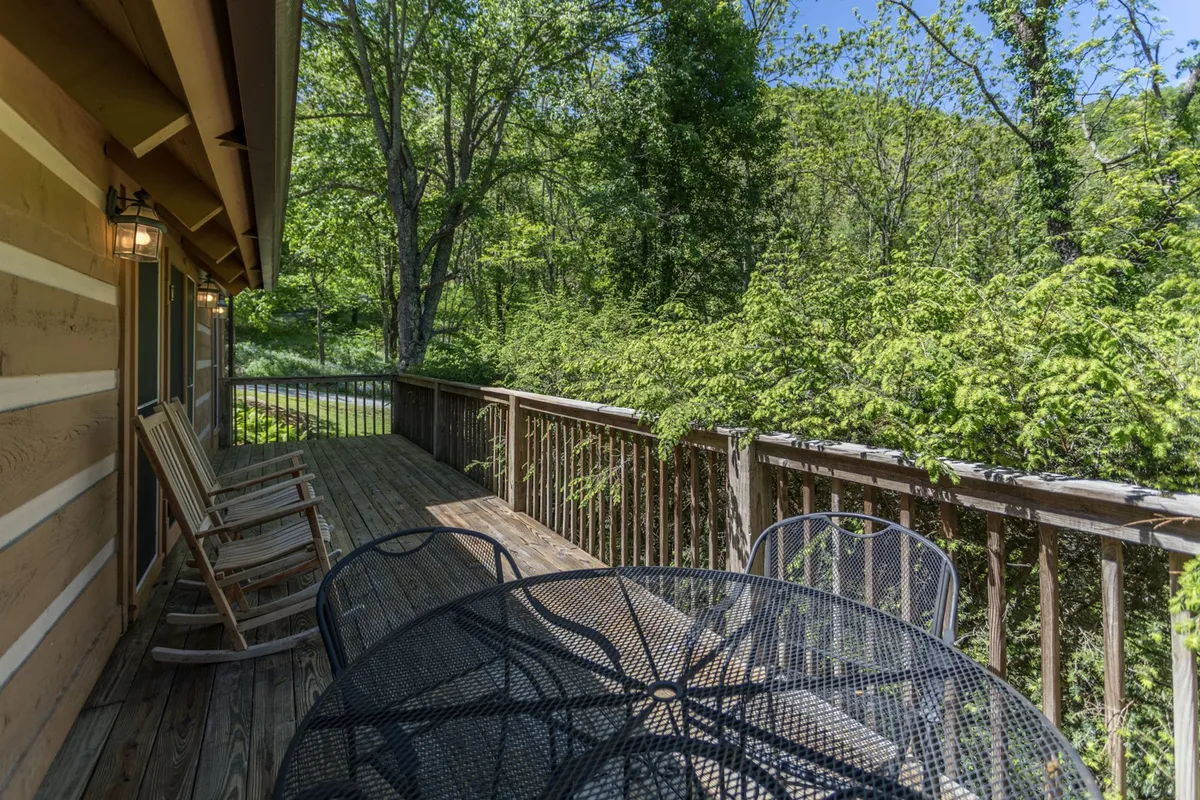 A peaceful wooden deck with classic rocking chairs, surrounded by lush greenery and trees. The deck offers a welcoming spot for morning coffee, quiet conversation, or stargazing at night. The metal table and comfortable seating create an inviting atmosphere for couples to relax and take in the mountain air.