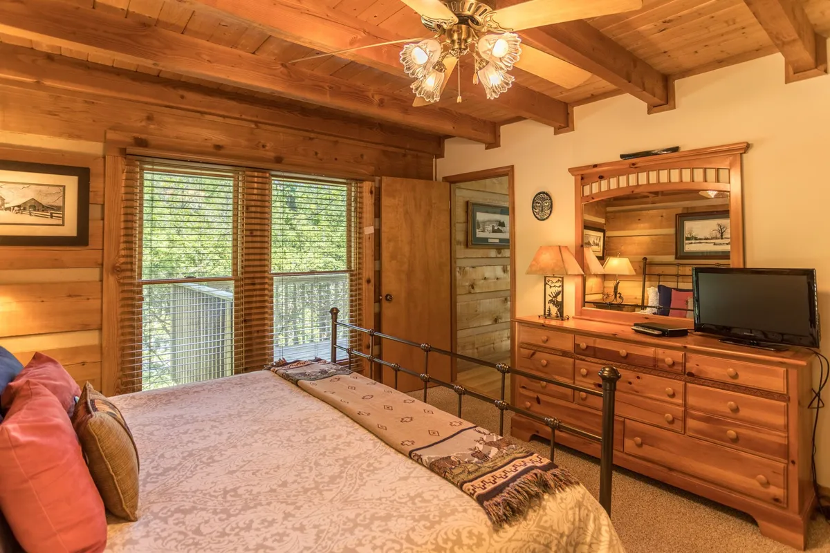 A welcoming bedroom with a bed adorned with decorative pillows, a wooden dresser and mirror, a window with blinds allowing natural light, and a ceiling fan overhead. The space feels private and comfortable, ideal for relaxation.