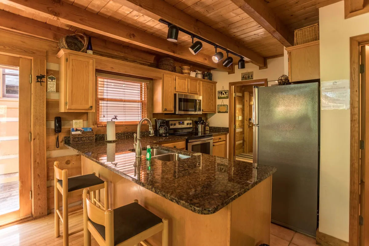 This image showcases a warm, inviting kitchen with a granite countertop, kitchen island, wooden cabinets, stainless steel appliances, and dining chairs. The kitchen is spacious and clearly designed for both meal preparation and socializing, suggesting ease and comfort for gatherings and home-cooked feasts.