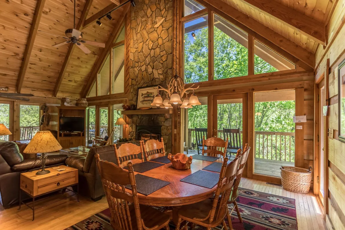 An interior view featuring a spacious dining area with a large rustic table positioned near the stone fireplace. Expansive windows provide outdoor views and illuminate the space, seamlessly connecting the living and dining zones.