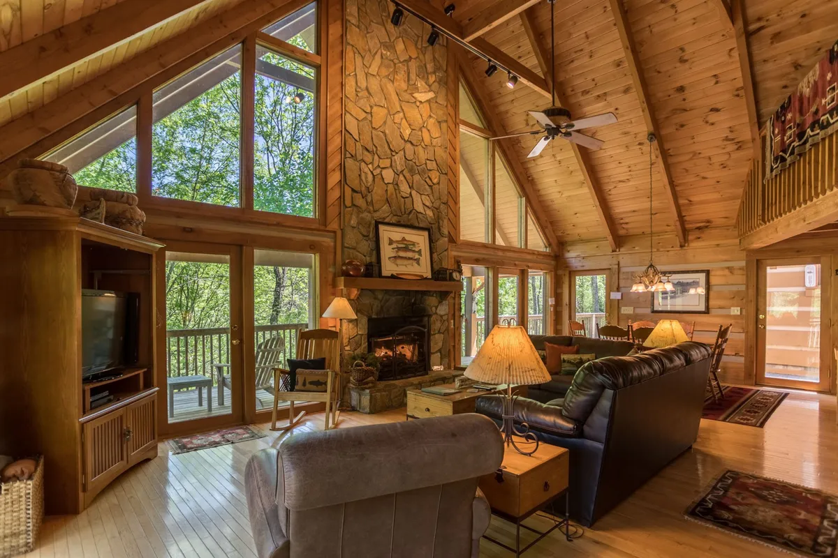 A welcoming living room centered around a stunning stone fireplace, surrounded by comfortable seating. Large windows invite natural light, accentuating the wooden ceiling and cozy atmosphere. The warm tones and arrangement encourage gathering and relaxation.