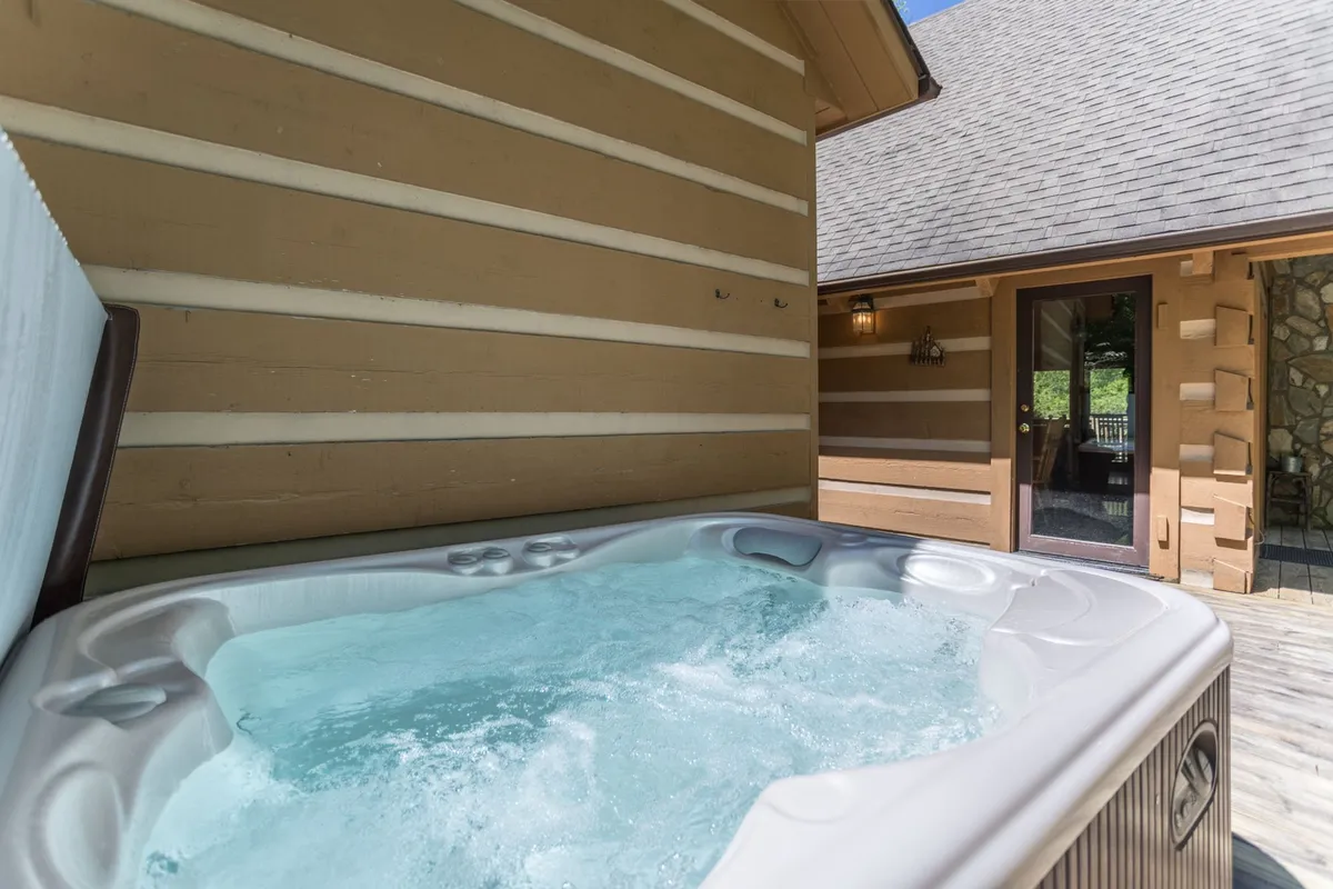 This image features an inviting hot tub nestled outside a wooden cabin, with the wooden exterior and a doorway that hints at the connection between indoor comfort and outdoor relaxation. The area is tranquil, offering a serene spot to unwind while being surrounded by natural elements.