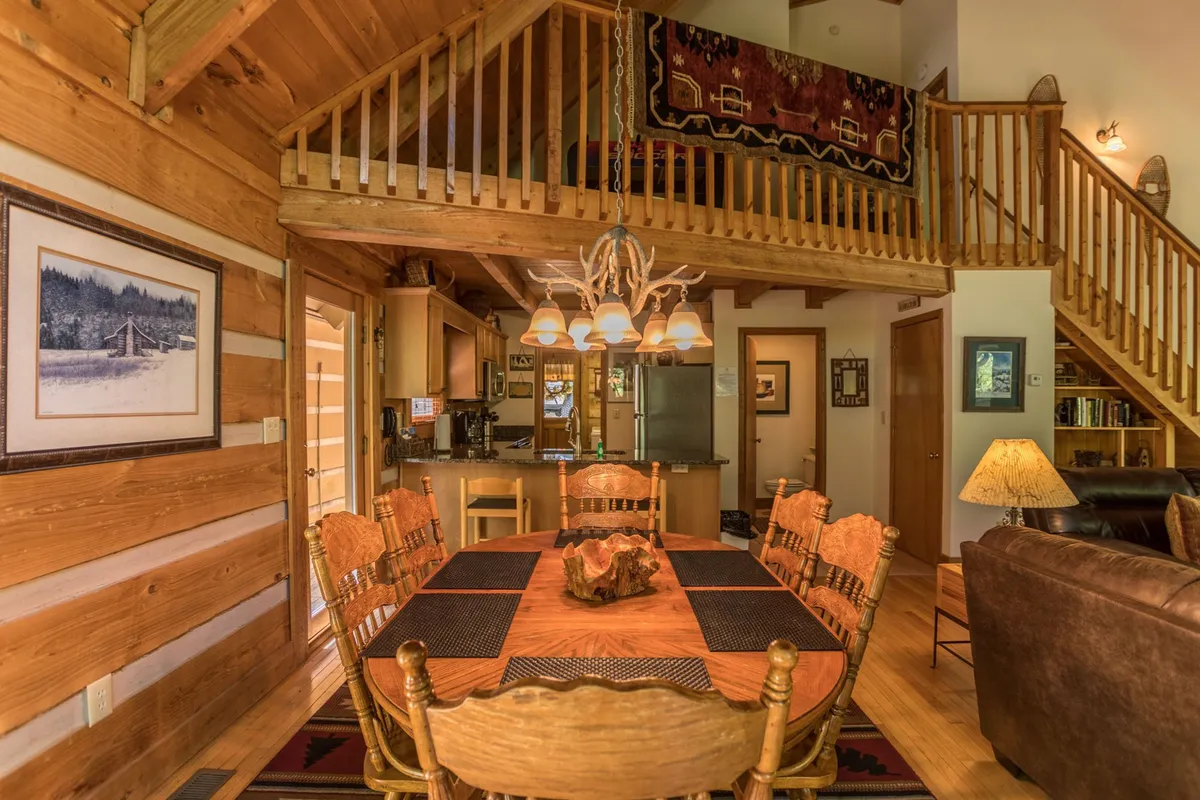 This scene features a welcoming dining area with a prominent dining table under a chandelier, a visible kitchen in the background, and a staircase adding architectural interest. The mood is warm and communal, perfect for shared meals, family dinners, and gatherings around the table.