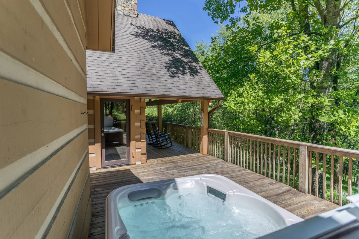 This exterior image features a spacious deck furnished with rocking chairs and a bubbling hot tub, surrounded by mature trees, with ample space for relaxing, socializing, and enjoying the outdoors. The setting conveys tranquility and connection to nature.