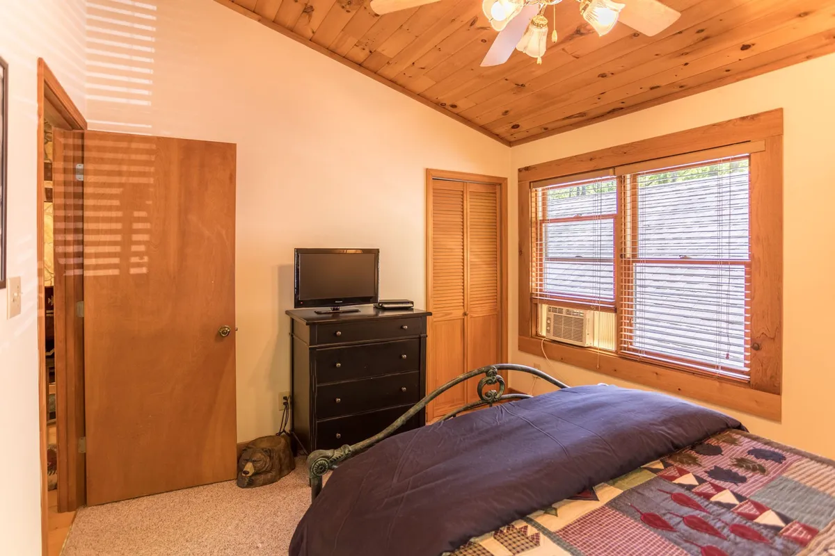 A cozy bedroom featuring a quilt-covered bed, a television for downtime, warm wooden furniture, and a window with blinds that allows natural light to brighten the space. The inviting setup suggests comfort for both people and pets, and the bed is styled in a way that evokes relaxation.