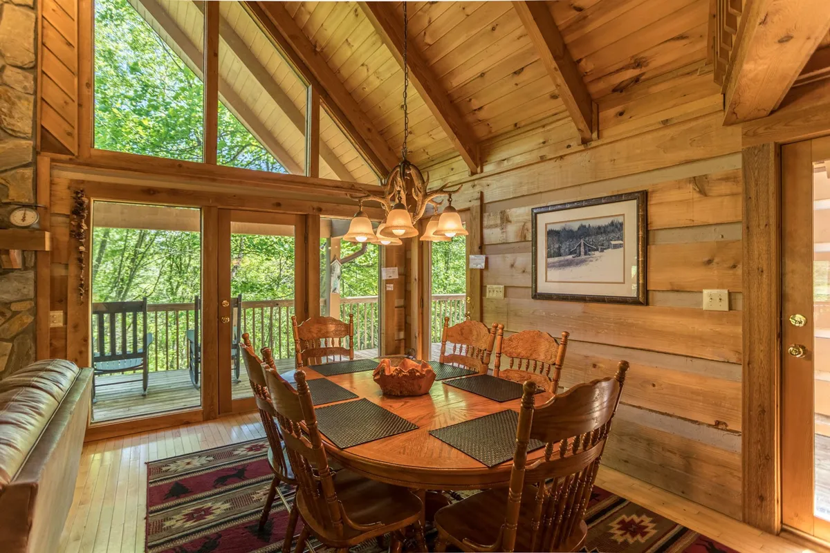 A large country-style dining table surrounded by wooden chairs, set next to expansive windows with views of the outdoors. The space features wooden beams and stone accents, evoking a rustic, inviting charm ideal for group meals.