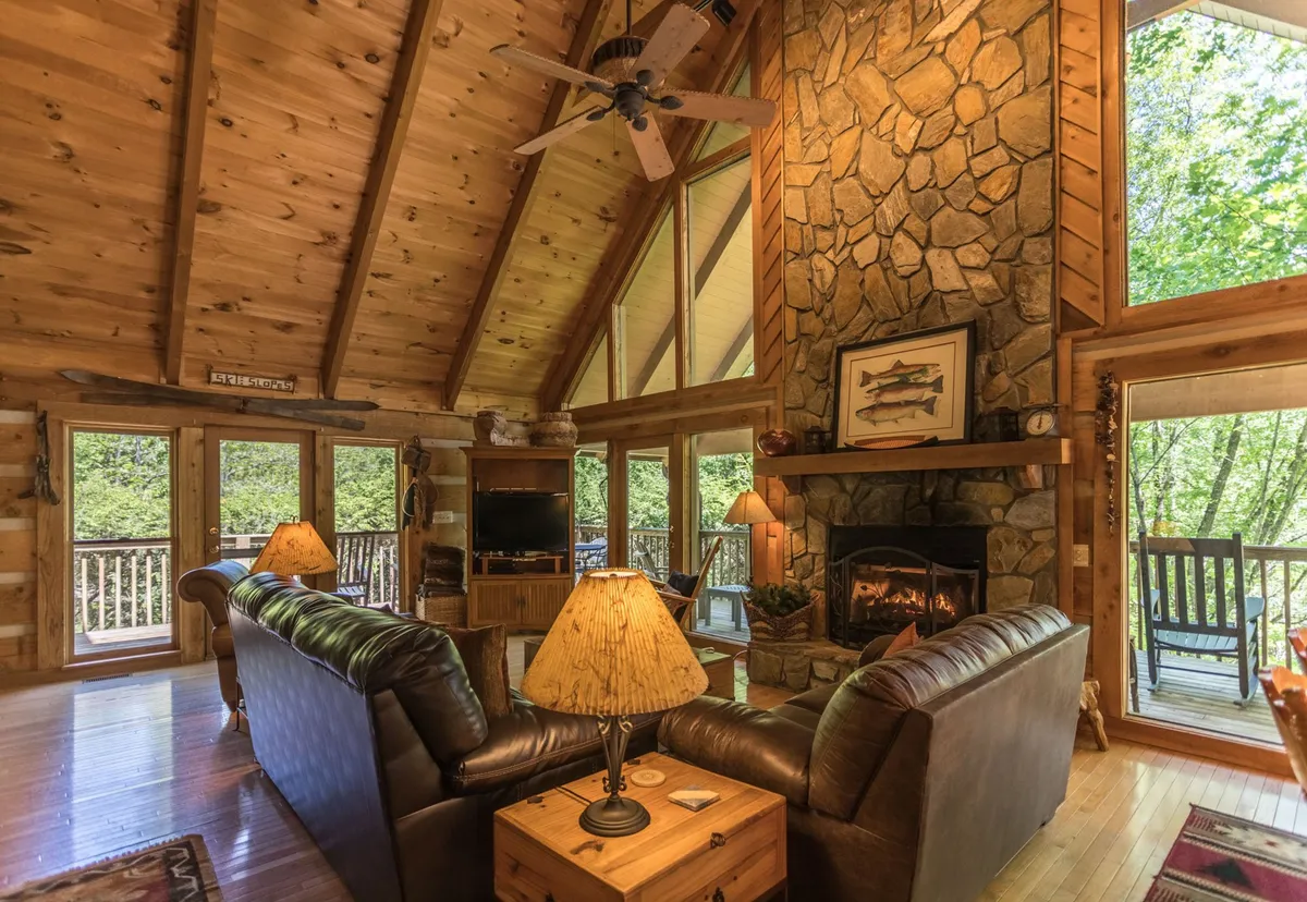 The interior hero image features a warm and welcoming living space with a striking stone fireplace as the centerpiece. Large windows bathe the room in natural light, highlighting the soaring wooden ceiling and rustic log cabin details. Plush leather sofas invite guests to relax, while the inviting ambiance suggests cozy gatherings, storytelling, and peaceful moments with mountain views.