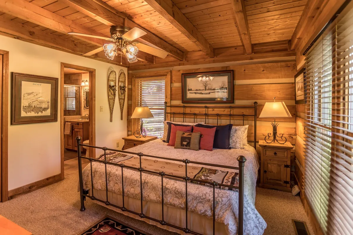 A cozy bedroom featuring a metal bed frame with decorative bedding, wooden ceiling and walls, soft table lamps casting a warm glow, and framed artwork that adds a local touch. The environment feels tranquil and intimate, ideal for restful nights.