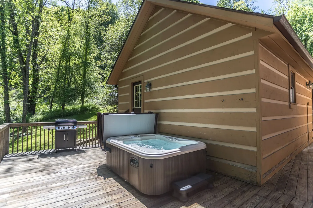 A tranquil exterior deck scene featuring a gas grill, hot tub, wooden deck, and lush trees. The setting is perfect for barbecues, alfresco dining, and relaxed socializing in the fresh mountain air.
