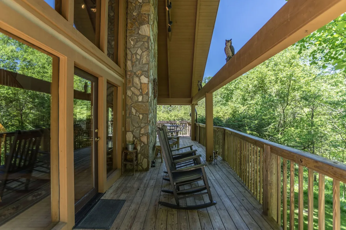 A serene porch scene with comfortable rocking chairs set against a stone wall and wooden railing, overlooking lush greenery and featuring an owl perched on the railing. The inviting arrangement encourages relaxation, wildlife watching, and enjoying the fresh mountain air.