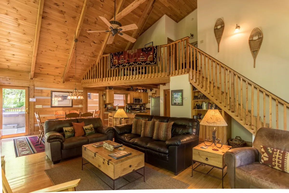A spacious open living area with high vaulted ceilings, a warm stone fireplace, leather sofas gathered around a wooden coffee table, a wooden staircase, and decorative lamps. The room is filled with natural light, creating a cozy, welcoming atmosphere perfect for groups.
